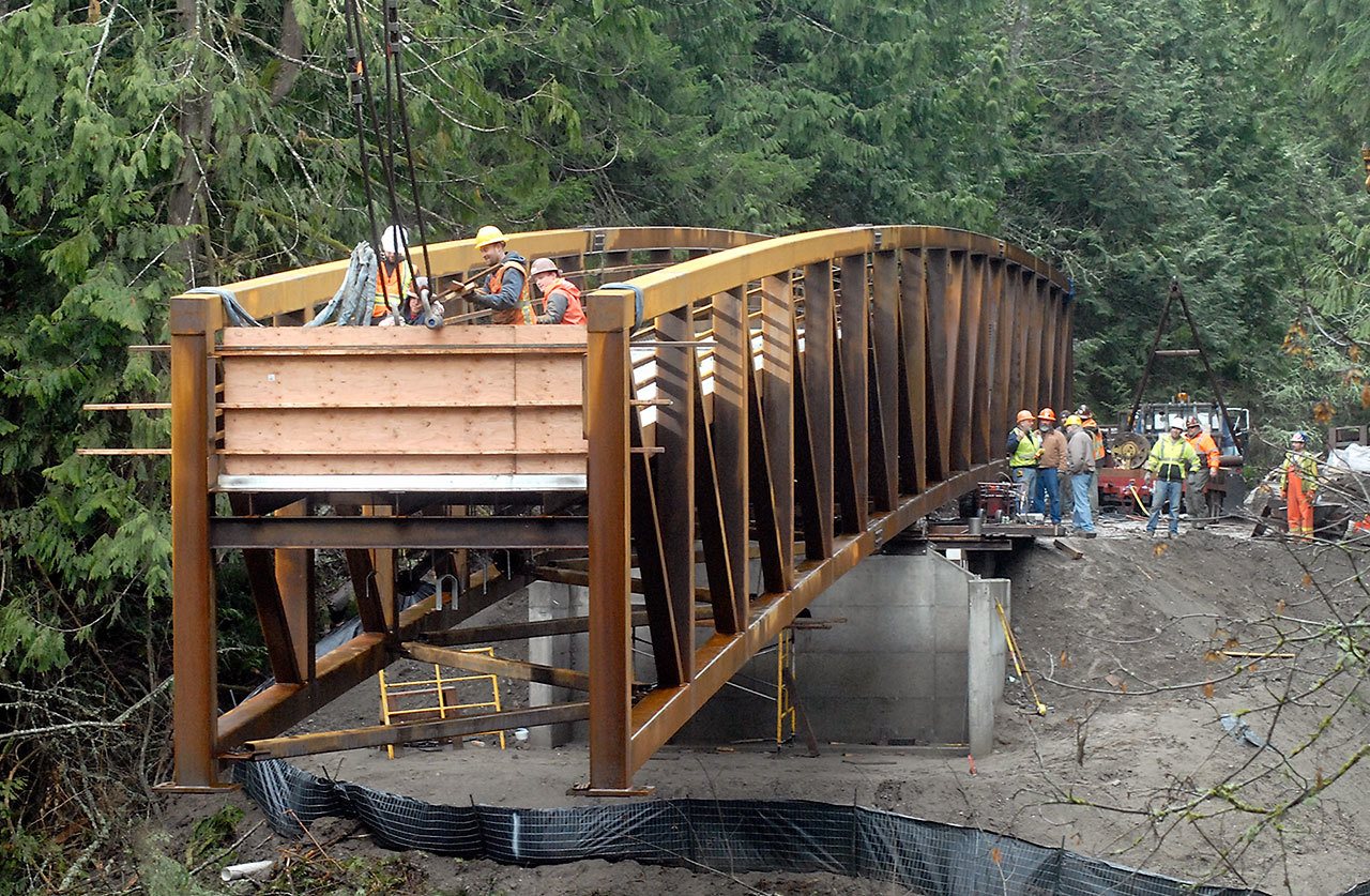 A rigging crew attaches the end of a 210-foot bridge span to crane cables as part of the structure hangs out over a small stream in Sequim Bay State Park east of Sequim on Thursday. The span replaces a former culvert crossing and will become a link in the Olympic Discovery Trail. (Keith Thorpe/Peninsula Daily News)