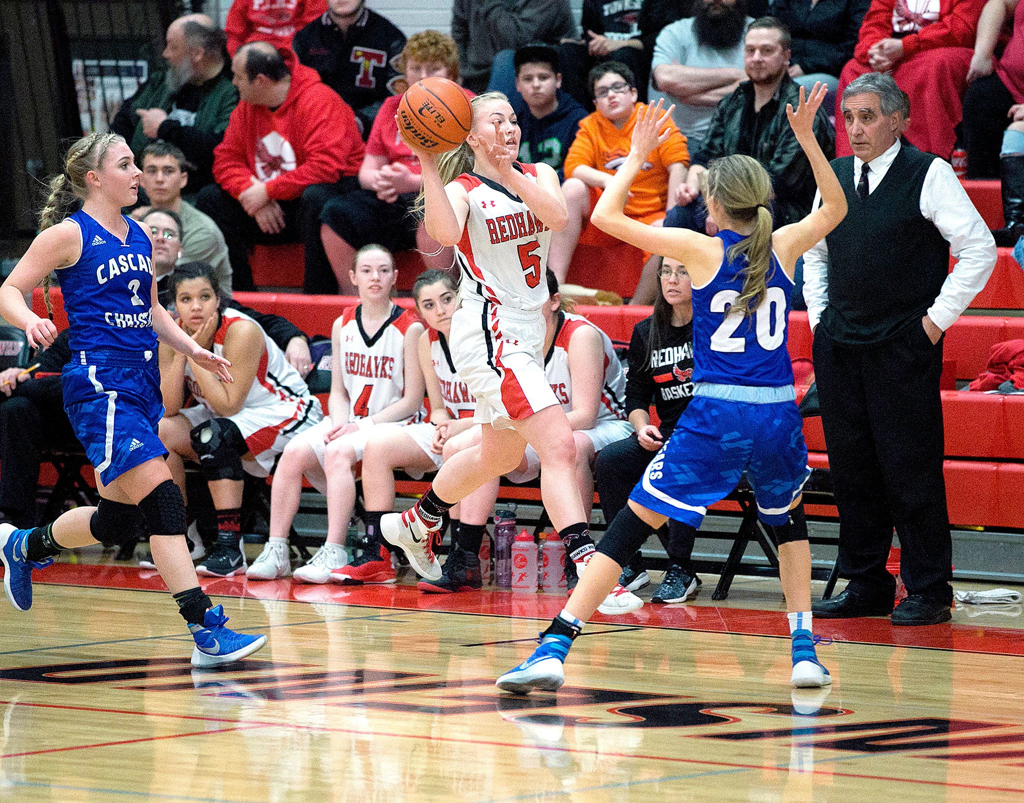 Steve Mullensky/for Peninsula Daily News Port Townsend’s Kaitlyn Meek looks to pass past Cascade Christian defenders as Redhawks’ head coach Scott Wilson looks on. Meek scored 18 points in the second half to rally the Redhawks from a huge deficit.