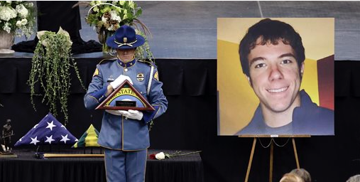 A State Patrol trooper holds a state flag to be presented to the family as he stands near a portrait of Thomas Zbyszewski at a memorial service for three firefighters killed in a wildfire. The Associated Press