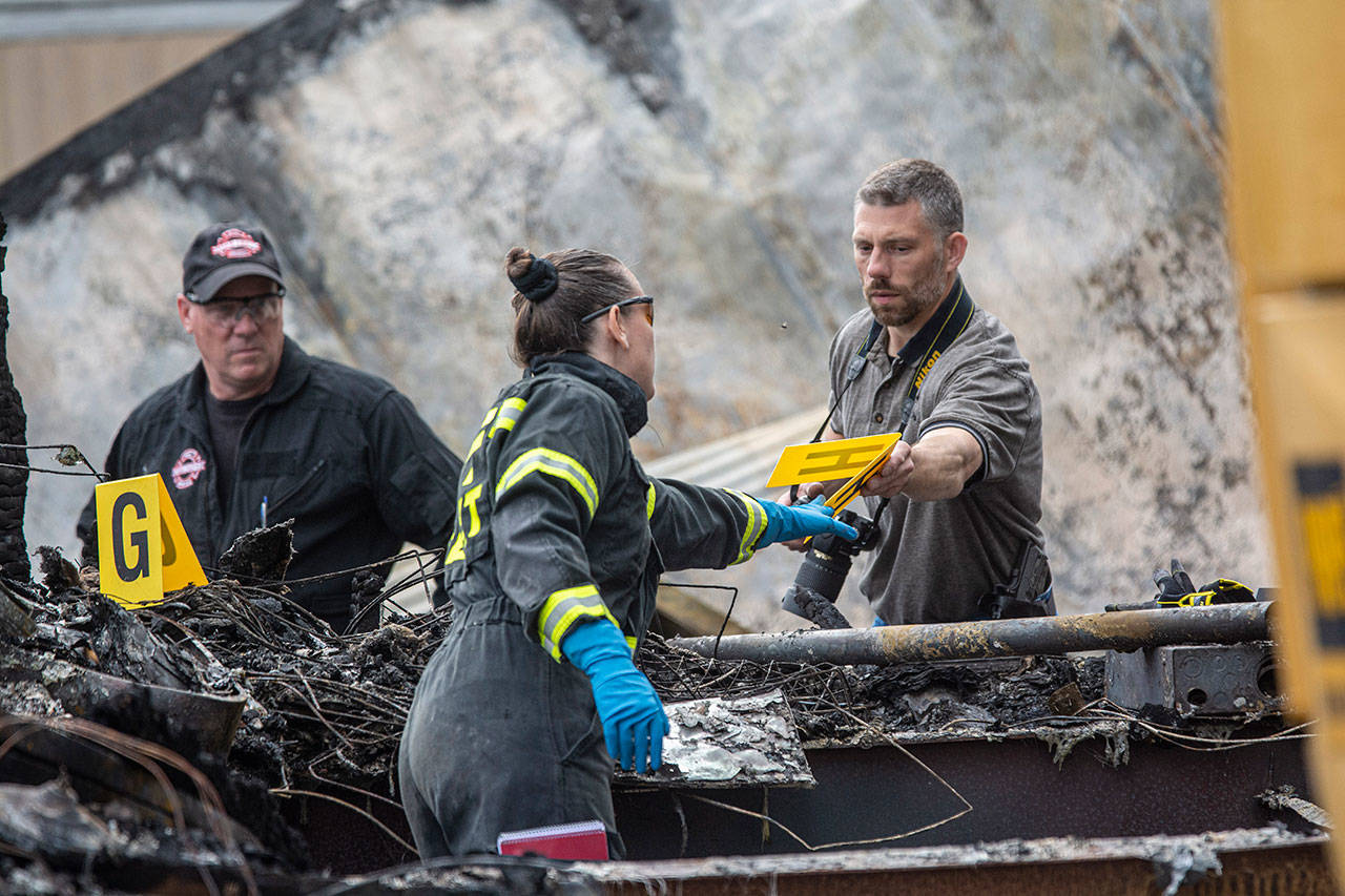 Clallam County Sheriff’s Office Detective Josh Ley, right, hands an evidence marker to an ATF agent as investigators comb through the scene of a fire where the bodies of a mother and three children were found. (Jesse Major/Peninsula Daily News)