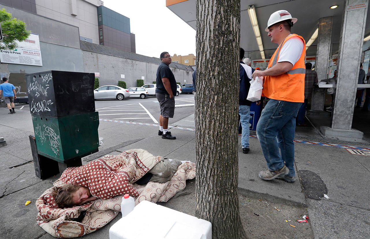 A man sleeps on the sidewalk as people behind line up to buy lunch at a Dicks Drive-In restaurant in Seattle in May 2018. An elected official in King County, where Seattle is located, wants to dedicate $1 million for one-way bus tickets to relocate homeless people who say they want to reconnect with family outside the region. (Elaine Thompson/The Associated Press)