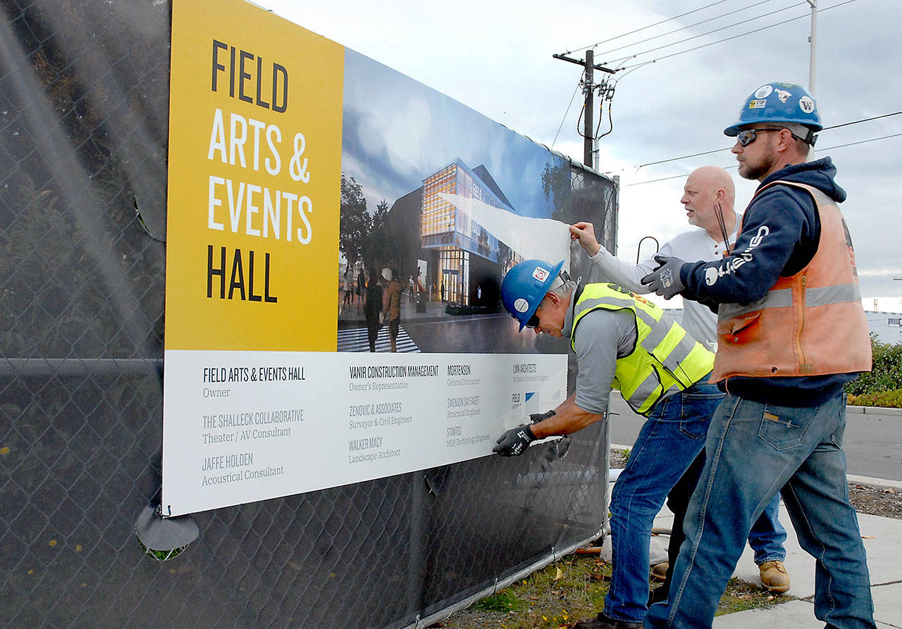 Chris Fidler, executive director of the Field Arts and Events Hall, center, gets assistance from Don Frostad, left, and Clayton Frohman of general contractor M.A. Mortenson Company as they peel back the protective cover from a project sign at the future site of the performing arts center at Front and Oak streets on the Port Angeles waterfront. (Keith Thorpe/Peninsula Daily News)