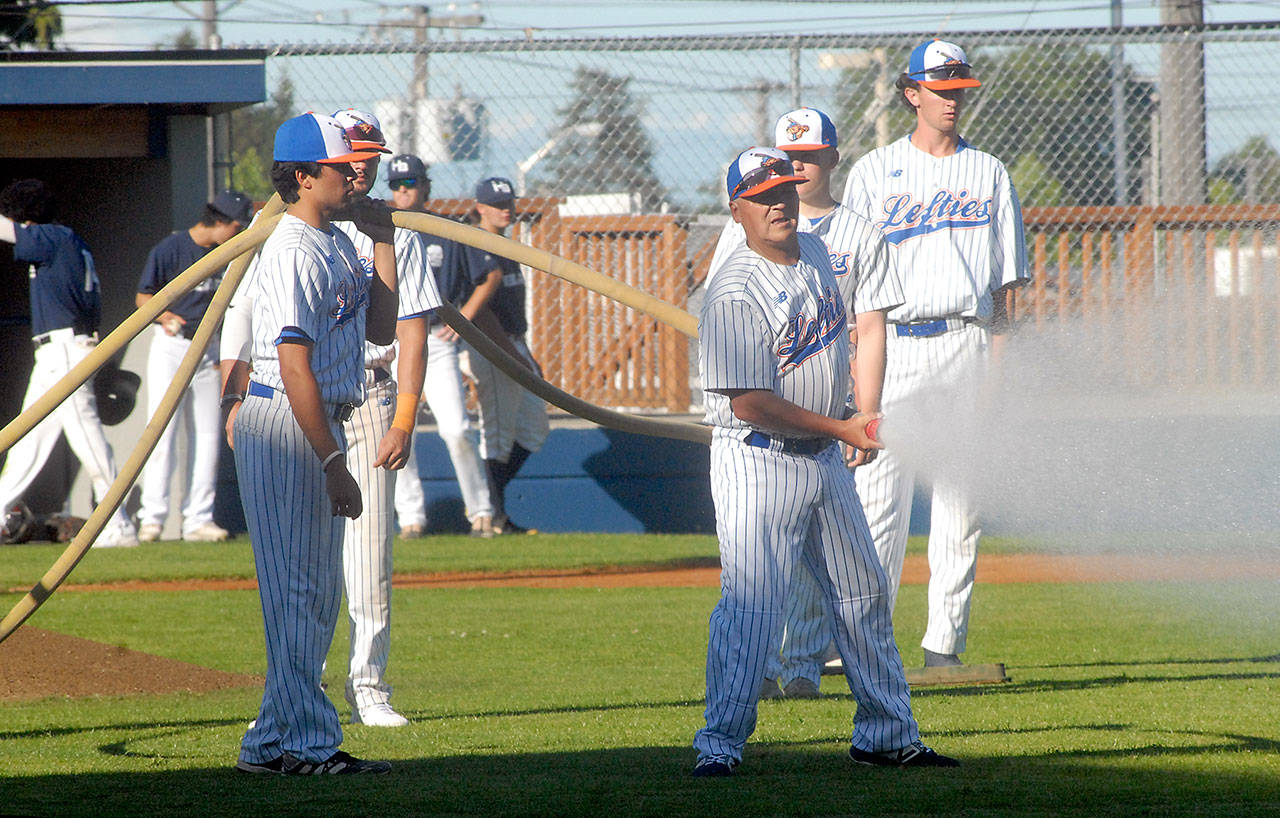 Members of the Port Angeles Lefties baseball team, including assistant coach Anthony Murillo, holding a hose, water down the infield prior to the start of Tuesday evening’s season opener against the Highline Bears at Port Angeles Civic Field. (Keith Thorpe/Peninsula Daily News)