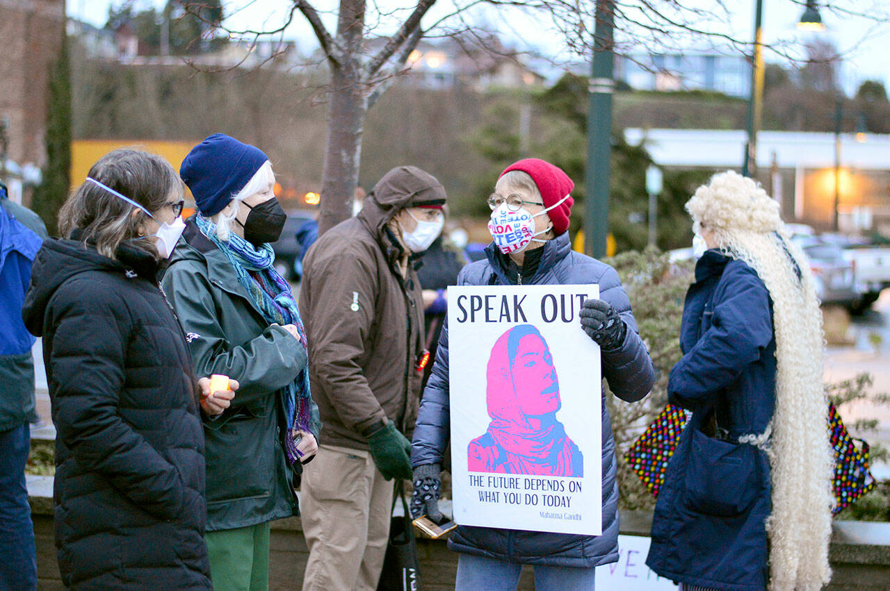 Debbi Steele of Port Townsend shows her sign to Mado Most, far left, and Annalee McConnell just before the candlelight vigil at Pope Marine Park in downtown Port Townsend on Thursday afternoon. About 100 people joined the League of Women Voters-Jefferson County vigil, marking the first anniversary of the insurrection at the U.S. Capitol. Earlier in the day, a vigil in Sequim drew at least 125 people. (Diane Urbani de la Paz/Peninsula Daily News)