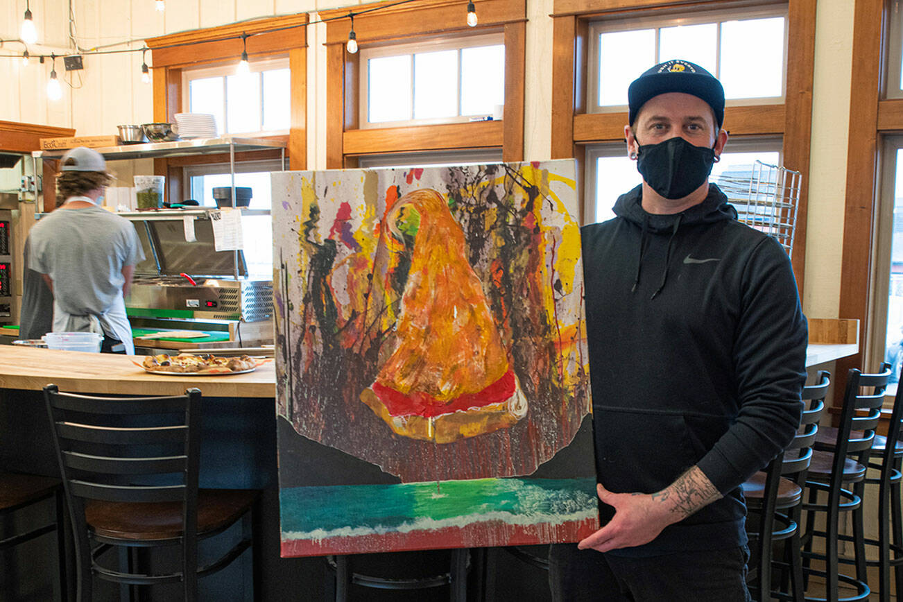 Larry White, district manager for Barhop Brewing & Artisan Pizza and artist, holds a painting for Barhop's new Sequim location.Behind him, kitchen manager Jeffrey Adams prepares pizzas for customers in the open kitchen. Emily Matthiessen/Olympic Peninsula News Group