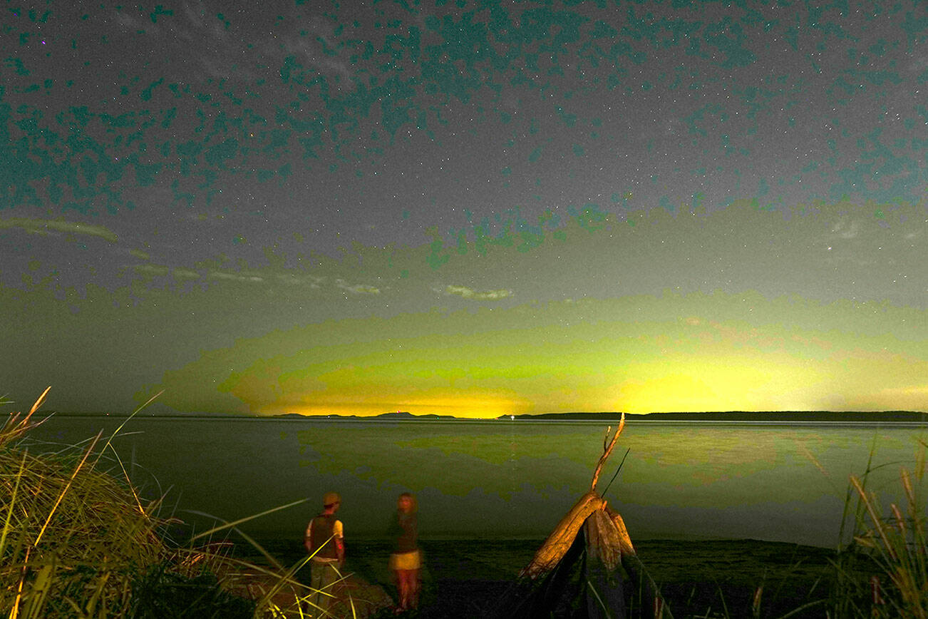 A couple watches as the stars twinkle above the colorful greens and purples from the Aurora Borealis as it shines above the light pollution over Whidbey Island on Wednesday night as seen from North Beach in Port Townsend. (Steve Mullensky/for Peninsula Daily News)