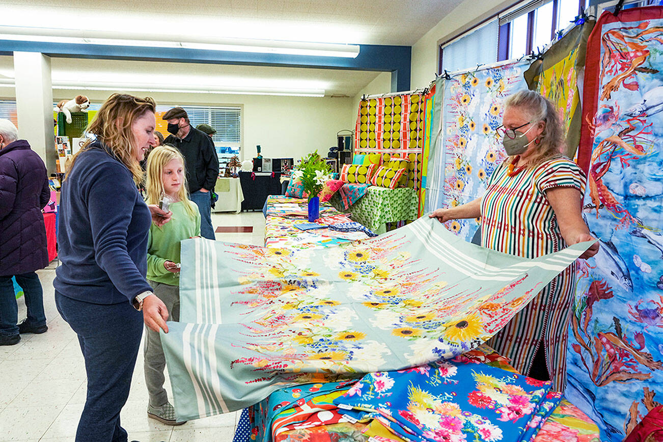 Steve Mullensky/for Peninsula Daily News

Martha Worthley, of Mim and Poppy Design, shows a tablecloth she designed to Alisa Martin and daughter Etta-Pearl, during the Port Townsend Arts Guild Arts and Crafts Fair on Friday at the Port Townsend Community Center in the Uptown neighborhood. The fair continues today from 10 a.m. to 5 p.m.