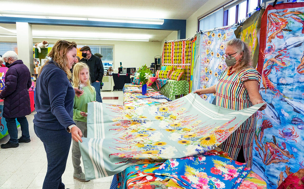 Martha Worthley, of Mim and Poppy Design, shows a tablecloth she designed to Alisa Martin and daughter Etta-Pearl, during the Port Townsend Arts Guild Arts and Crafts Fair on Friday at the Port Townsend Community Center in the Uptown neighborhood. The fair continues today from 10 a.m. to 5 p.m. (Steve Mullensky/for Peninsula Daily News)