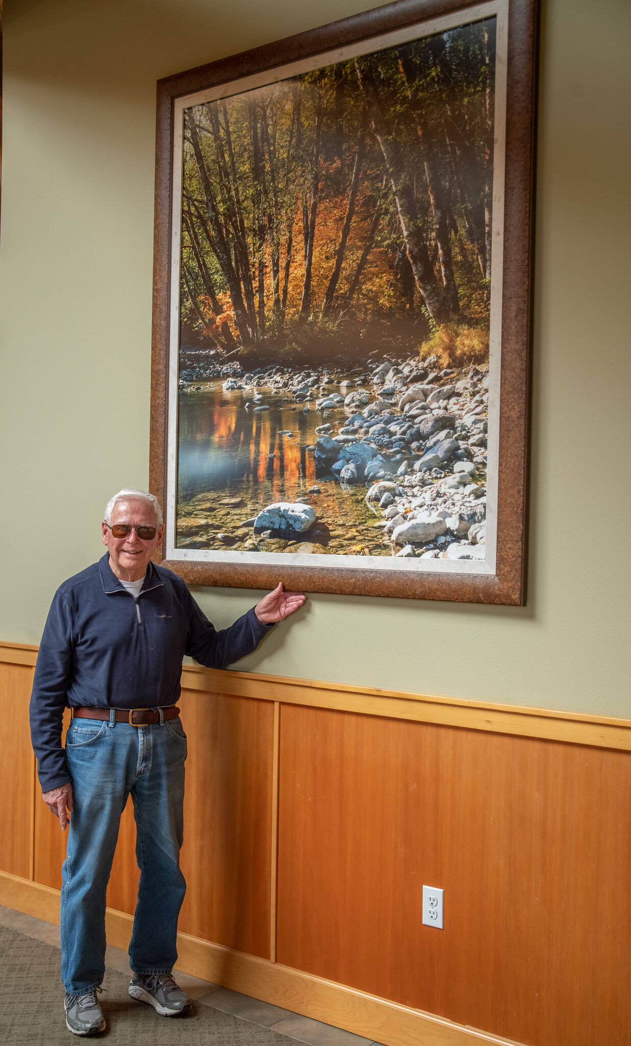 Emily Matthiessen / Olympic Peninsula News Group
Photographer Ross Hamilton stands with one of the 35 prints of his 4-by-5-inch negatives that are displayed in the hallways of the Lodge at Sherwood Village. Hamilton says that each of his photographs has a story behind it; this one was a brief moment in time when the light overwhelmed him with its beauty coming through the trees on the Dosewallips River. “The joy of seeing something really beautiful is what makes photography so special,” he says.