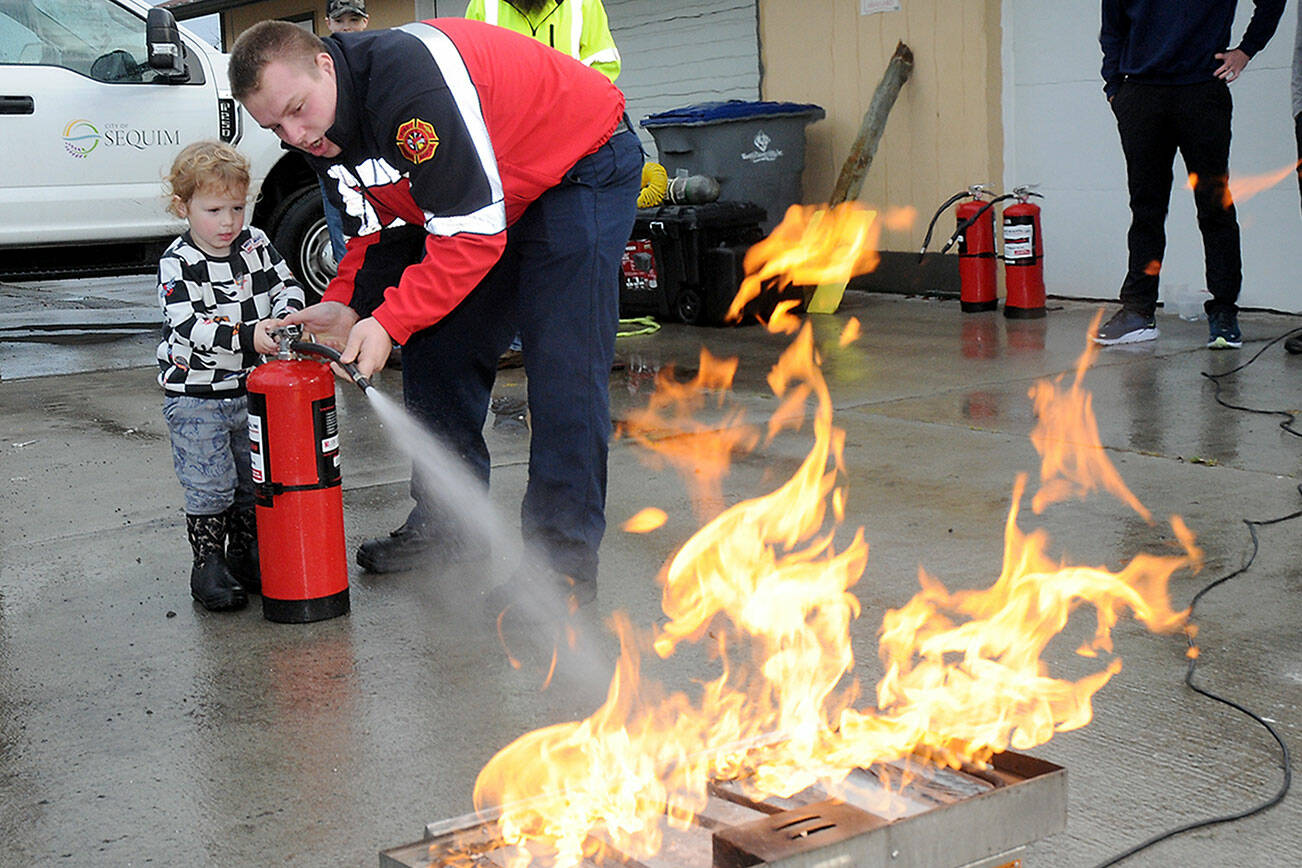 Beau Rossi, 2, of Sequim gets assistance from Clallam County Fire District 3 firefighter/paramedic Hayden Pyle with extinguishing an intentional propane fire during Saturday’s Public Safety and Information Fair at Carrie Blake Park in Sequim. The event, organized by Fire District 3 and the Community Emergency Response Team, featured a variety of exhibits and displays from numerous public safety and community service agencies, as well as workshops and children’s activities. (Keith Thorpe/Peninsula Daily News)