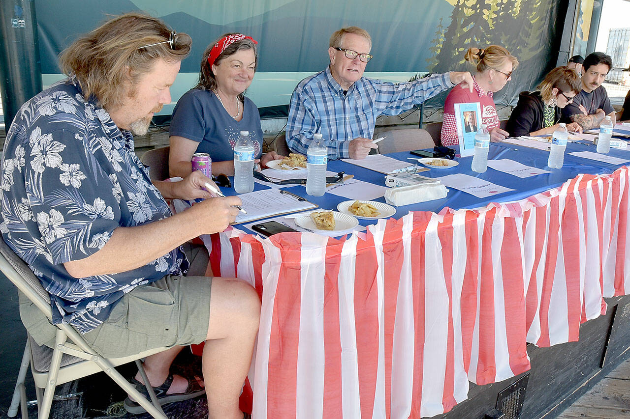 Pie contest judges, from left, Jeff Tocher, Laurel Hargis and Clallam County Commissioner Randy Johnson, along with event sponsor Country Aire Natural Foods representatives Kristina Fallon, Katie Meyers and Caeron Alarcon, make their choices during Thursday’s annual Independence Day apple pie contest at Port Angeles City Pier. A total of 26 pies were entered into the contest with first-place honors going to Xaven McCarty of Seattle, Elisabeth Pennell of Mukilteo in second and Selena Reach of Port Angeles taking third. The contest was dedicated the late legacy judge and former Peninsula Daily News publisher John Brewer, represented by a photo in front of an empty chair. (Keith Thorpe/Peninsula Daily News)