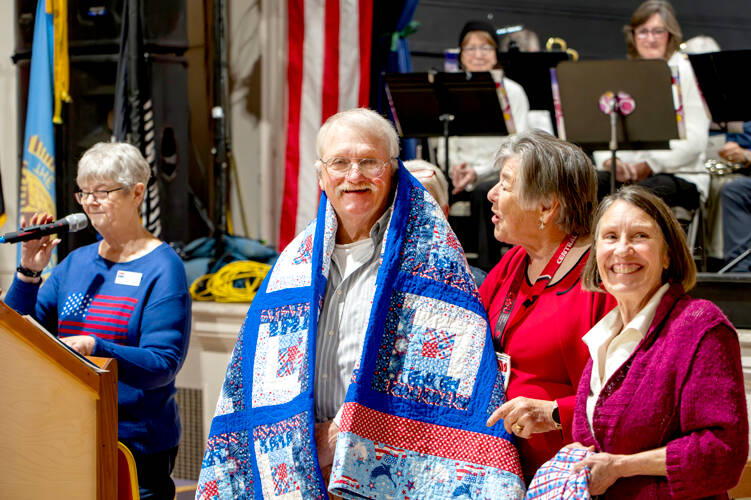 Former Marine Joseph Schwann of Port Townsend smiles as he receives a Quilt of Valor from Susan Travis, right, and another member of Quilts of Valor during the Veterans Day event at the American Legion Marvin G. Shields Memorial Post 26 in Port Townsend on Monday. Group leader Kathey Bates, left, was the emcee of the event. (Steve Mullensky/for Peninsula Daily News)