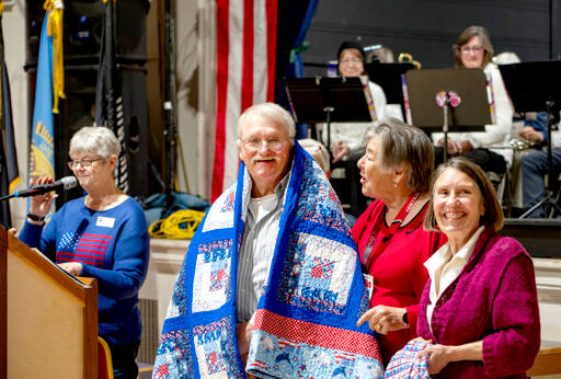 Former Marine Joseph Schwann of Port Townsend smiles as he receives a Quilt of Valor from Susan Travis, right, and another member of Quilts of Valor during the Veterans Day event at the American Legion Marvin G. Shields Memorial Post 26 in Port Townsend on Monday. Group leader Kathey Bates, left, was the emcee of the event. (Steve Mullensky/for Peninsula Daily News)