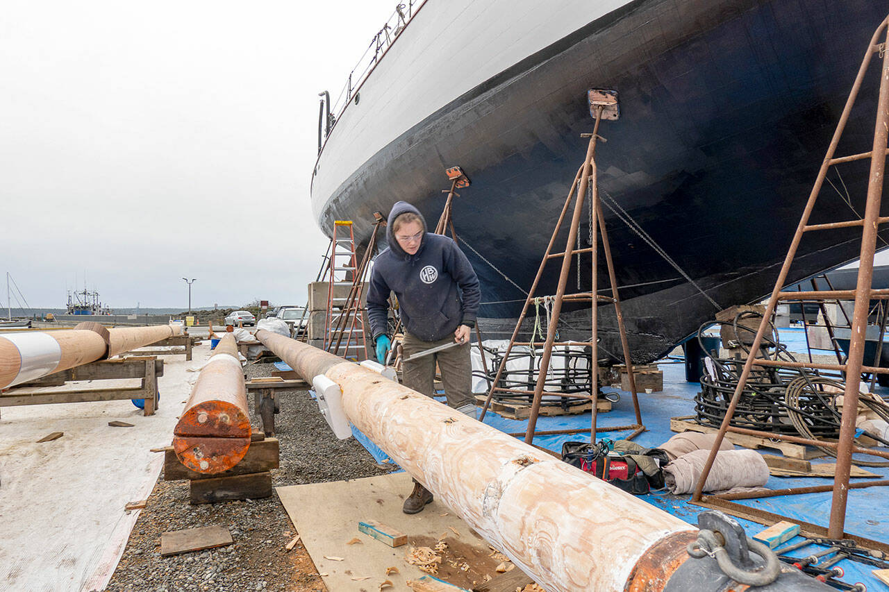 Zoe Hewitt, a shipwright at Haven Boatworks, uses a draw knife to shape the foremast on the 112-year-old historic schooner Adventuress, background, that is resting on the hard at the Port Townsend Marina. A new main mast, left, and the fore mast will be stepped next week, weather permitting. (Steve Mullensky/for Peninsula Daily News)