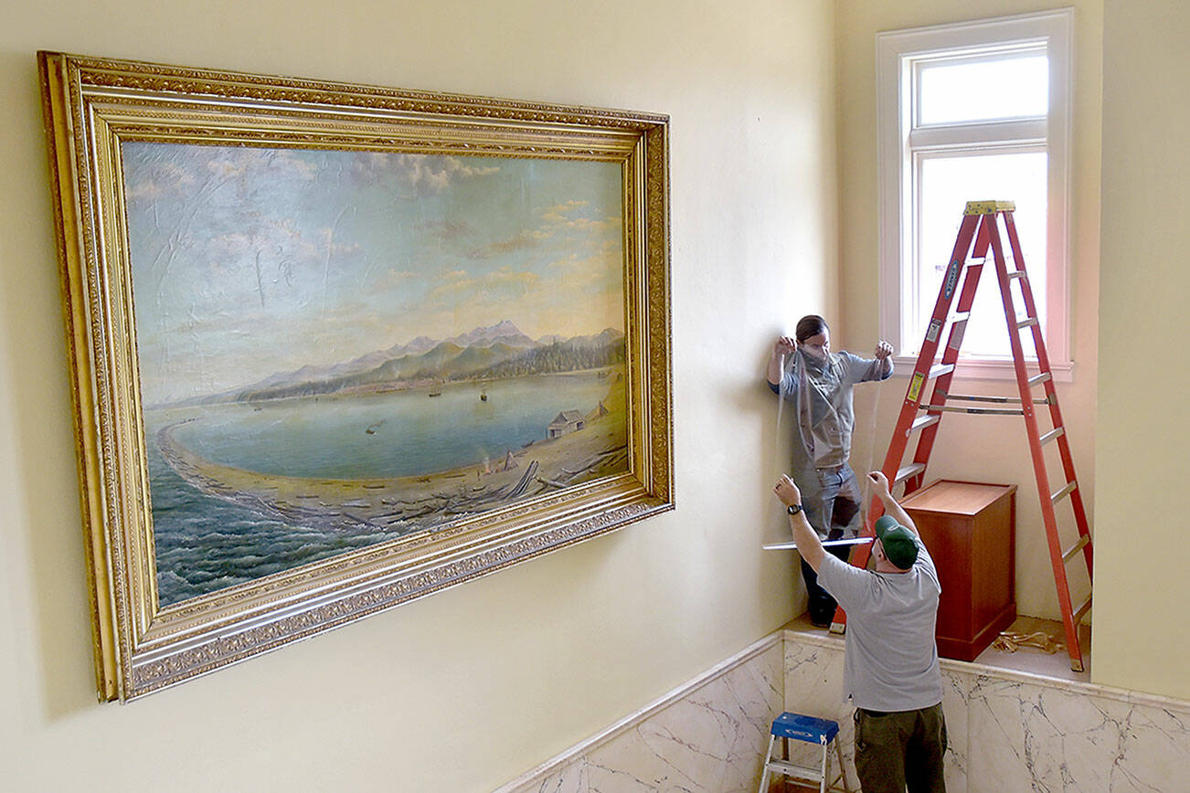 David Brownell, executive director of the North Olympic History Center, top, takes a piece of ultraviolet-filtering window tinting from Ralph Parsons, Clallam County maintenance worker, in an effort on Tuesday to protect historic paintings on the stairway of the section of the county courthouse, including an 1890s depiction of Port Angeles Harbor by artist John Gustaf Kalling. The history center is working with the county to preserve the stairway artworks by adding the window coatings to reduce damage from sunlight and installing an electronic UV monitor to track potentially harmful rays. (Keith Thorpe/Peninsula Daily News)