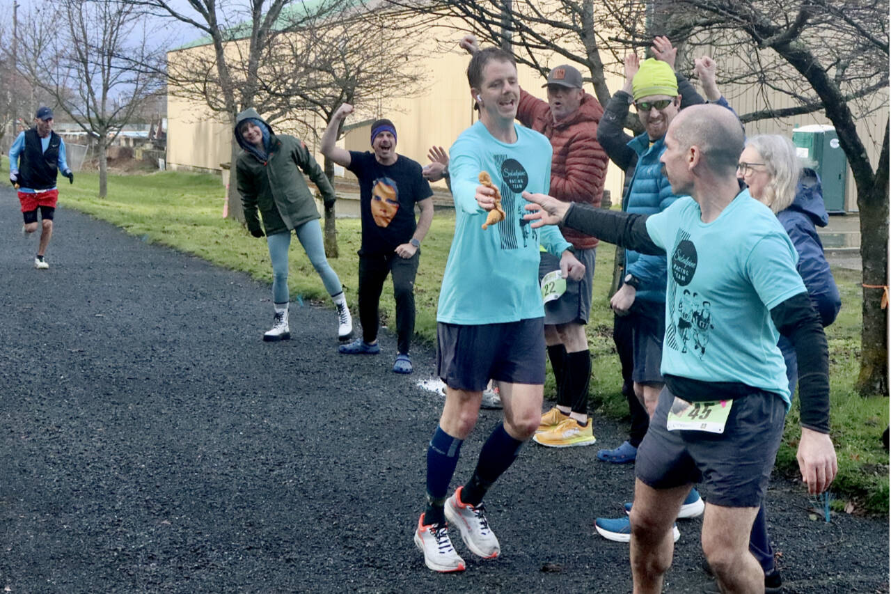 Chris Hartman of the Sub Alpine Tees Team hands off the crocheted yellow banana slug baton to teammate Jon Cummings. Fellow teammates cheer on the runners at the Port Angeles Yacht Club Club exchange station Saturday. (Dave Logan/for Peninsula Daily News)