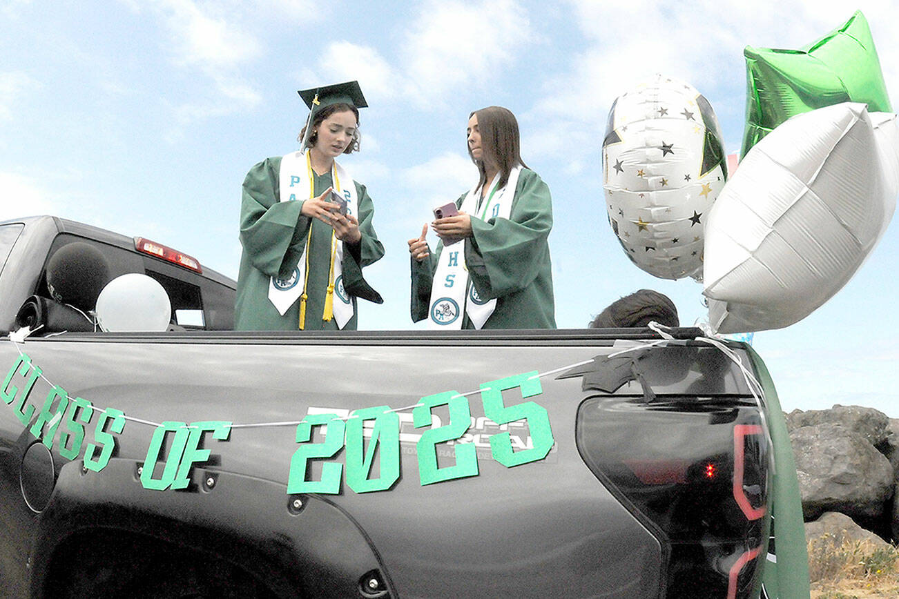 Port Angeles High School twin graduates Kaylie Mast, left, and Kendall Mast, communicate with their friends by text while waiting for the start of the 2025 graduation parade on Friday. The parade began at Ediz Hook and culminated with a formal ceremony Friday evening at Port Angeles Civic Field. (Keith Thorpe/Peninsula Daily News)
