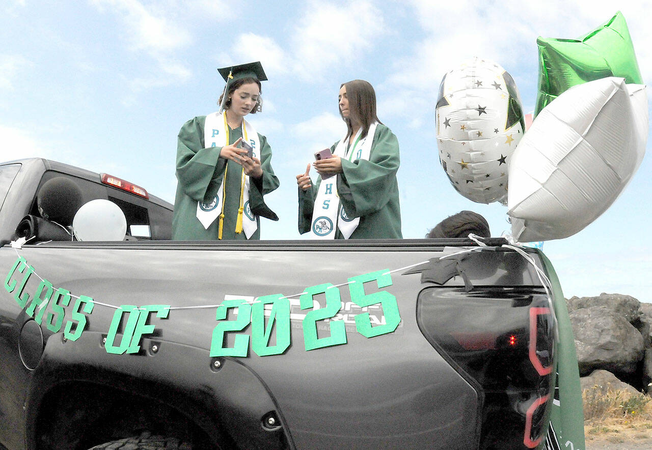 Port Angeles High School twin graduates Kaylie Mast, left, and Kendall Mast, communicate with their friends by text while waiting for the start of the 2025 graduation parade on Friday. The parade began at Ediz Hook and culminated with a formal ceremony Friday evening at Port Angeles Civic Field. (Keith Thorpe/Peninsula Daily News)