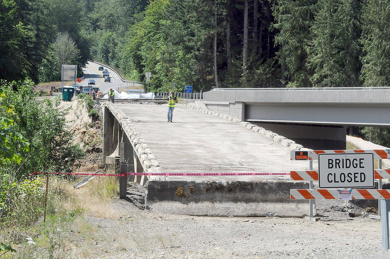 Construction workers stand on what remains of the old U.S. 101 bridge over the Elwha River on Wednesday as the aging structure is dismantled. The old bridge, built in 1926, was in danger of washout when the river beneath changed course and engineers discovered the bridge piers were built on gravel instead of bedrock, leading to construction of a new bridge, at right, which was opened to traffic in 2024. The old bridge was to remain in place until a fish-spawning window, which runs from mid-July until the end of August. (KEITH THORPE/PENINSULA DAILY NEWS)