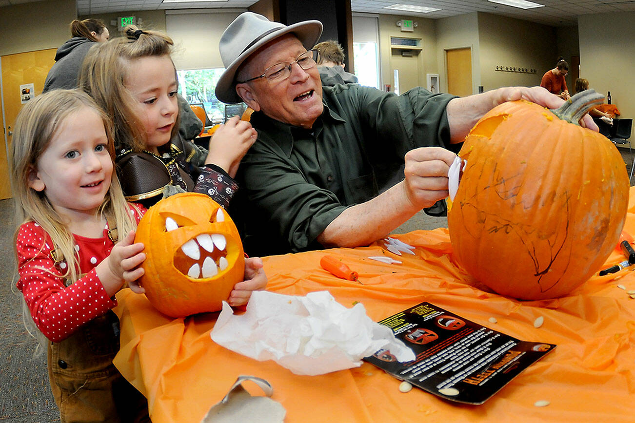 Port Angeles siblings Gwendolyn McCord, 4, left, and Brân McCord, 6, assist their grandfather, Glenn Jones, with creating a jack-o-lantern during a pumpkin-carving session on Saturday at the Port Angeles Public Library. About 40 children and their parents took part in the spooky Halloween activity. (Keith Thorpe/Peninsula Daily News)