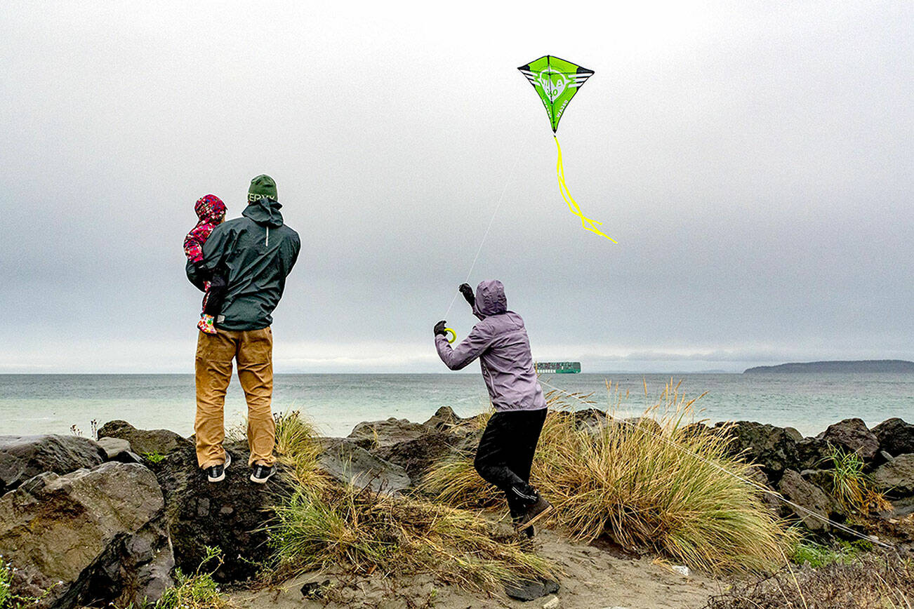Mara, 3, held by her father Mike Karas, watch as her mother, Molly, launches a kite into the 15 mph winds off the rocks at the Point Wilson Lighthouse on Sunday at Fort Worden State Park. (Steve Mullensky/for Peninsula Daily News)