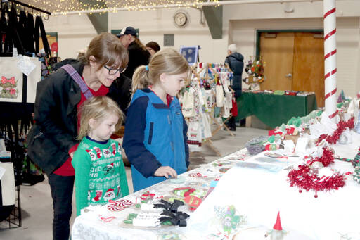 Christine Leaver and her two daughters, Sullivan, 5, and Avery, 9, look over many Christmas ideas on Friday during the annual Christmas Cottage at the Vern Burton Community Center in Port Angeles. More than 34 different vendors fill the gym with holiday spirit. The event will continue from 9 a.m. to 5 p.m. today and Sunday. (Dave Logan/for Peninsula Daily News)