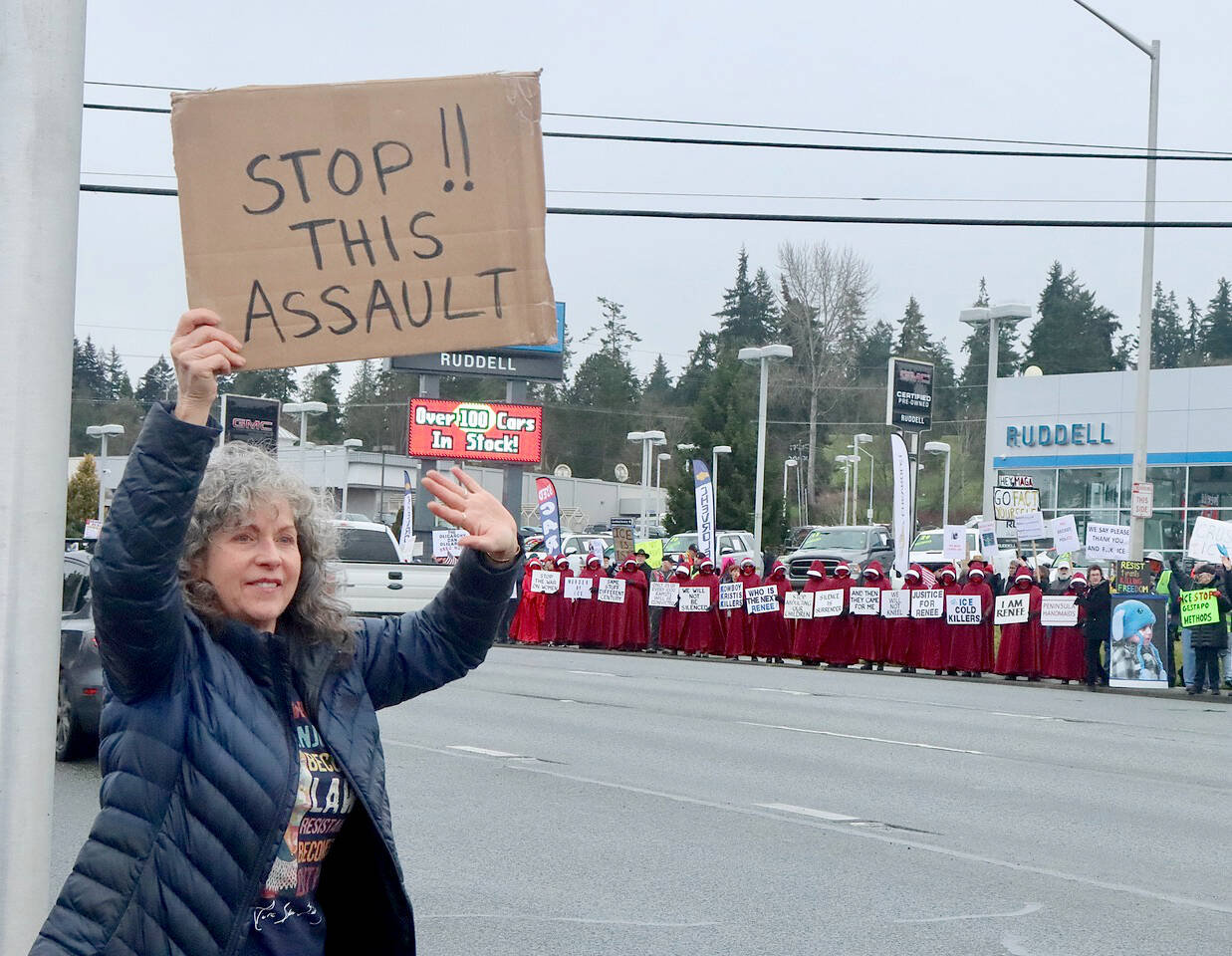 Wendy Rae Johnson waves to cars on the north side of U.S. Highway 101 in Port Angeles on Saturday during a demonstration against U.S. Immigration and Customs Enforcement operations in Minnesota. On the other side of the highway is the Peninsula Handmaids in red robes and hoods. (Dave Logan/for Peninsula Daily News)